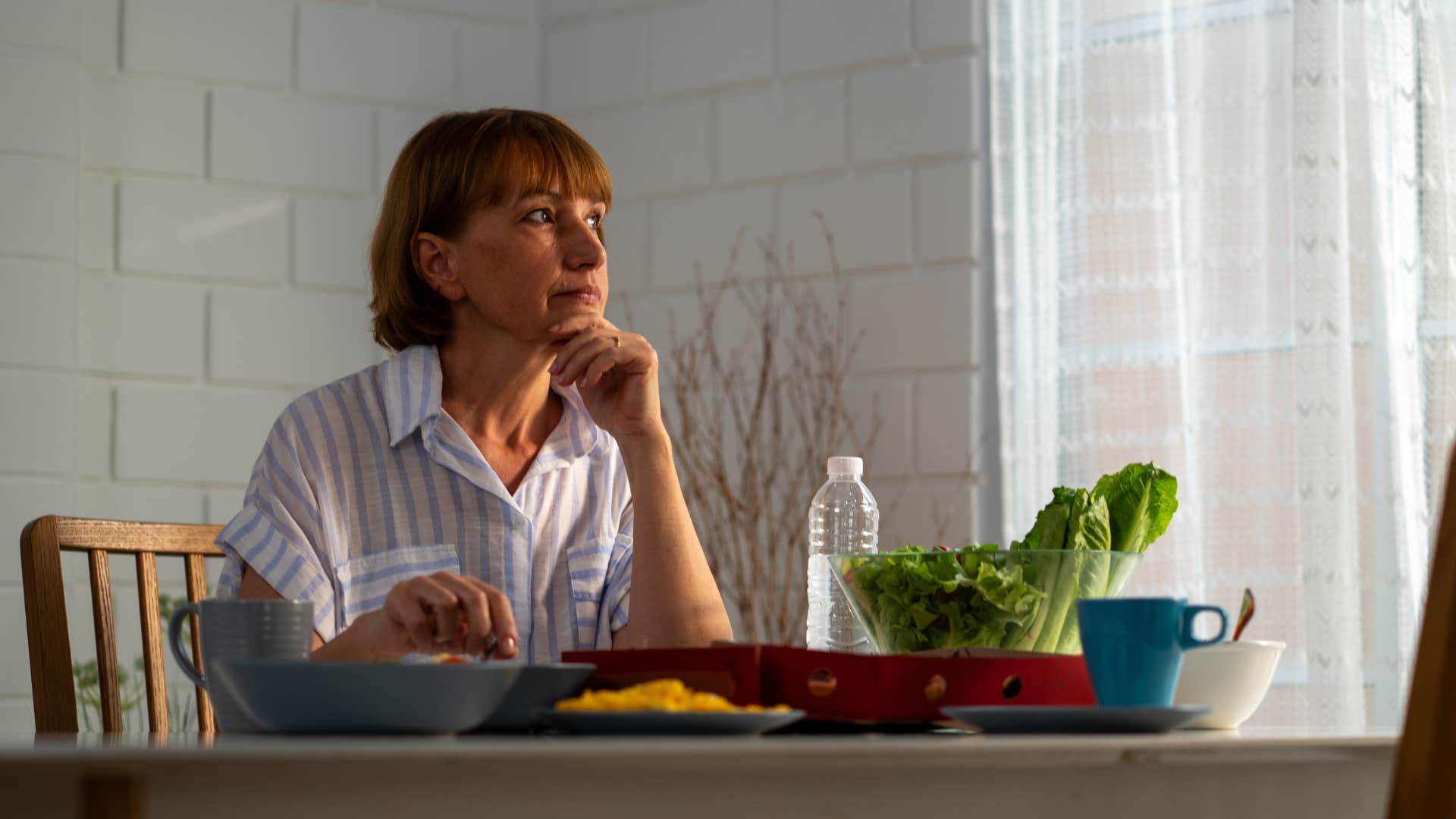 Sad woman sitting at her dining room table looking out a window.