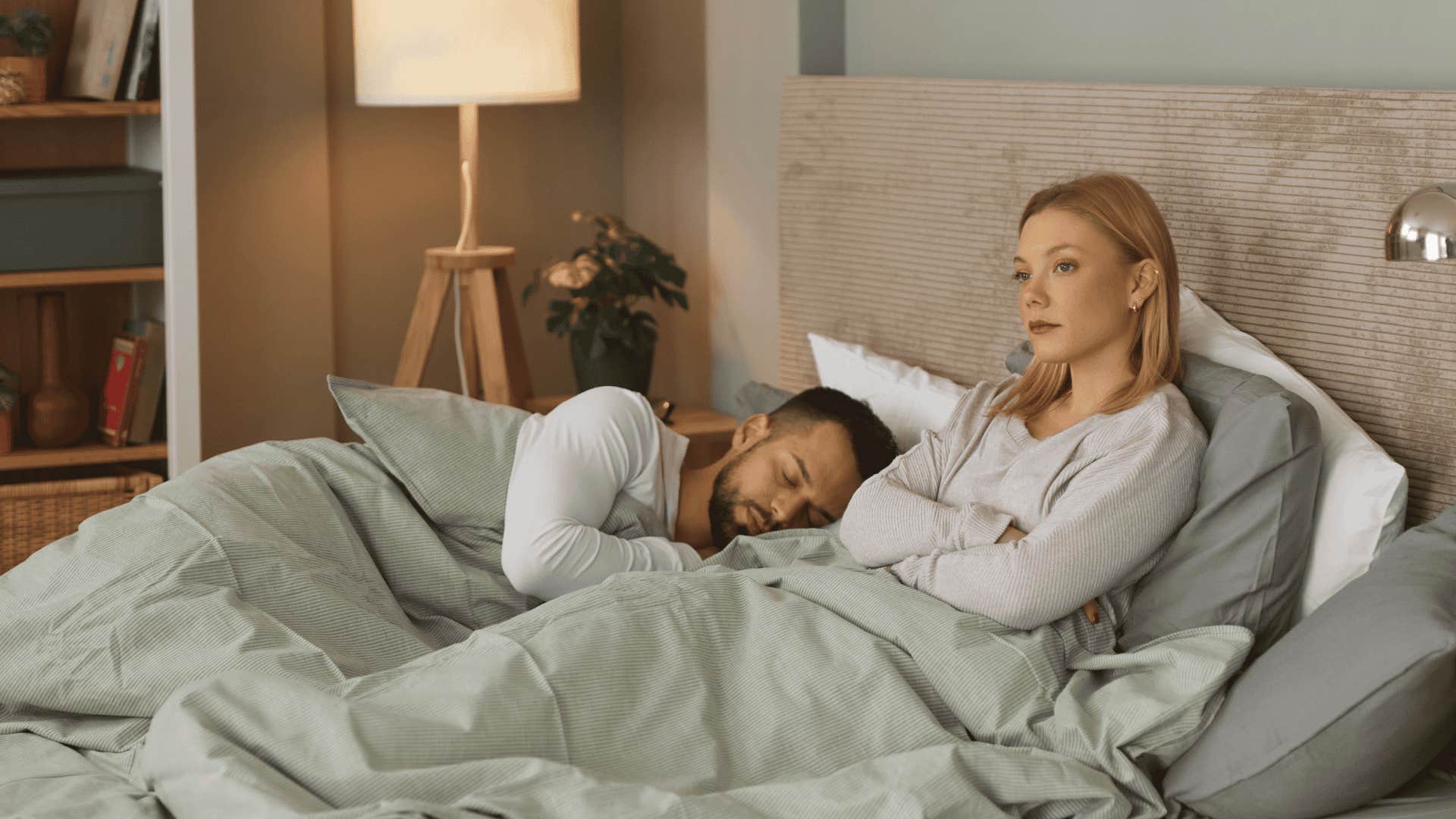 Upset woman sitting on bed with partner sleeping in background