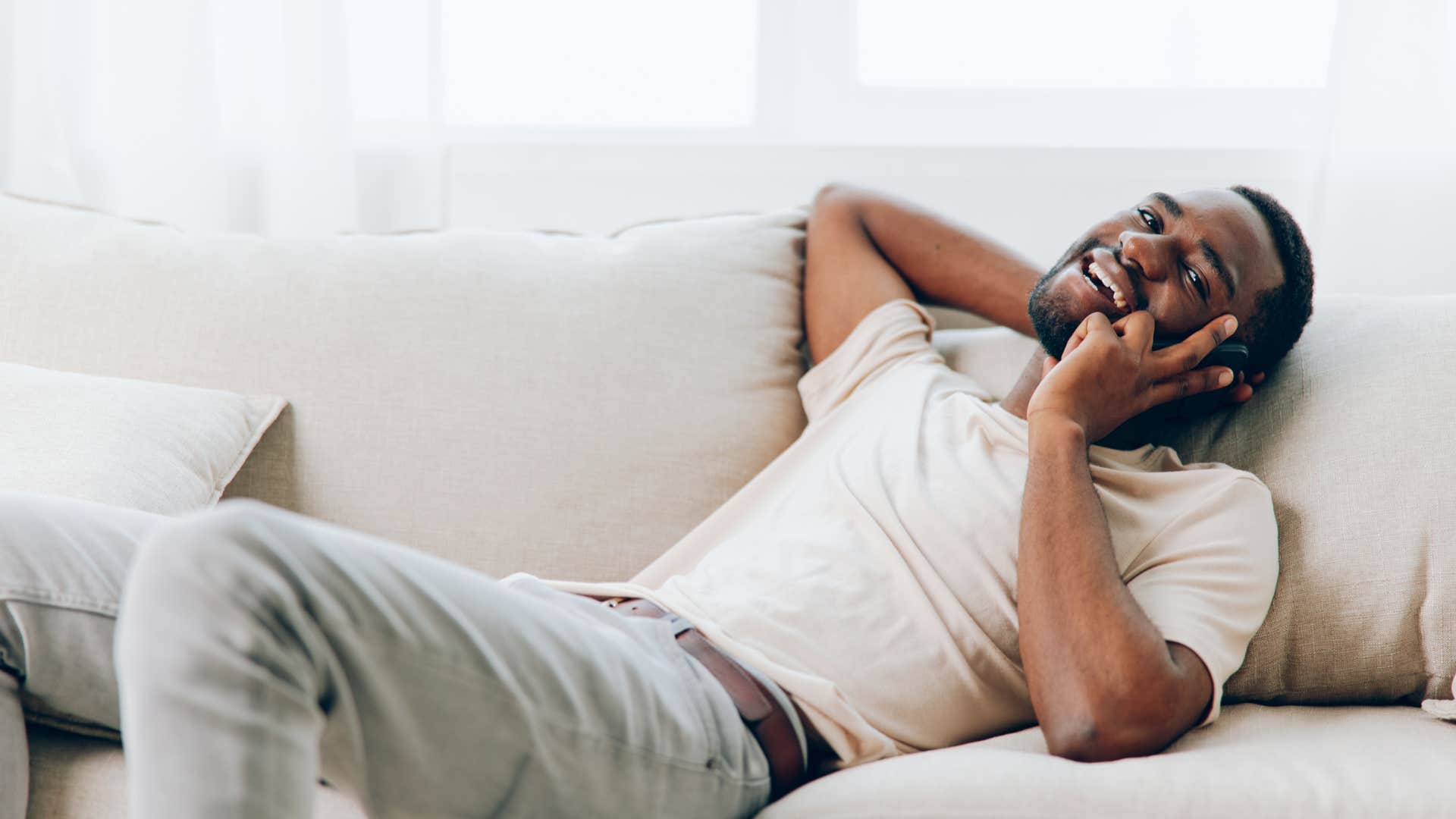 man laying on white couch talking on phone