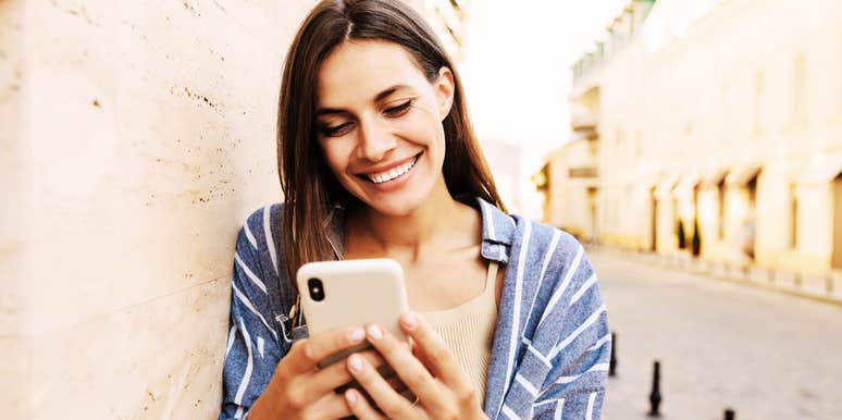 Woman leaning on wall smiling big, texting.