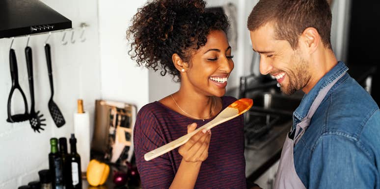 Couple cooking Italian food in kitchen, laughing taste testing red sauce. 