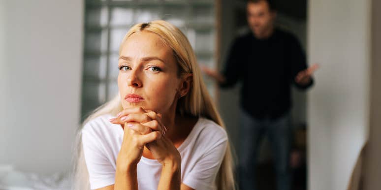 Stoic woman sitting on bed, upset husband trying to control her.