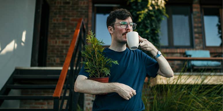 man drinking coffee in front of his house holding a small pine tree