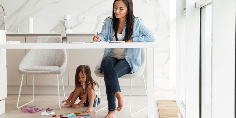 A mom with a smile on her face works while her child is Playing under the desk