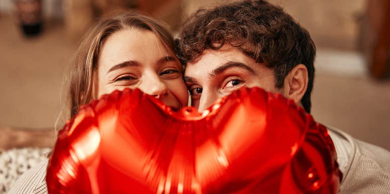 Young couple in love holding a heart-shaped balloon, hiding behind it while sitting on the sofa