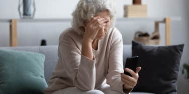 upset middle aged woman looking at phone screen sitting on couch at home