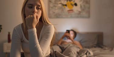 woman sitting on bed while partner sneakily texting on phone