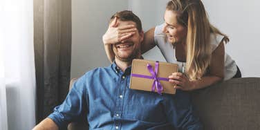 wife covering husband's eyes while handing him wrapped present