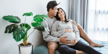 man and pregnant woman embrace each other while sitting by window at home