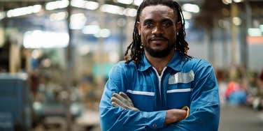 man in a blue uniform stands in a factory with his arms crossed.