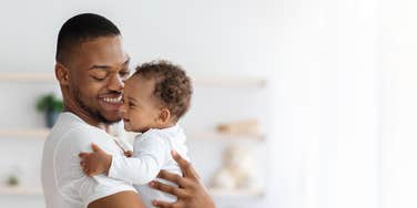 Portrait Of Happy African American Father With Infant Baby In Arms Standing Near Window At Home