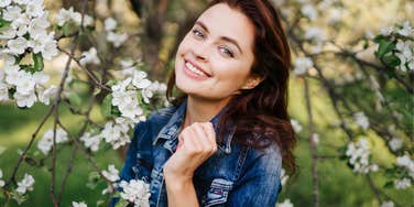 Young smiling woman in jeans jacket standing near blooming spring tree. 