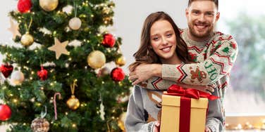 couple holding a gift standing next to a Christmas tree