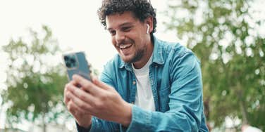 young bearded man in denim shirt sitting in wireless headphones making video call on mobile phone on busy street modern city background.