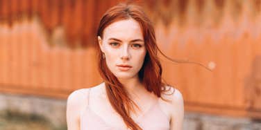 Close up portrait of young woman with long red hair and freckles posing outdoors on copper background. 