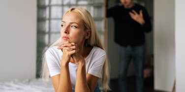 upset woman sitting on bed while man stands behind her trying to console her