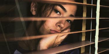 woman looking sadly through window blinds