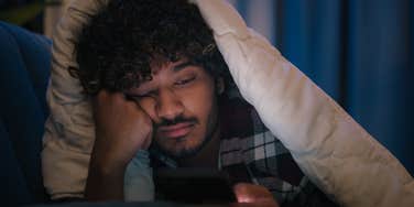 Sad man laying in bed scrolling on his phone.