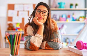teacher woman wearing sweater and glasses sits at desk looking frustrated and annoyed with crossed arms.